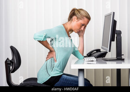 Una donna si siede alla scrivania e ha dolori alla schiena. Foto simbolico per la posizione di destra al lavoro in ufficio., Eine Frau sitzt am Schreibtisch und Foto Stock