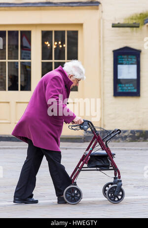 Donna anziana spingendo un aiuto per la mobilità a piedi lungo il telaio condividono lo spazio area a Cirencester, Gloucestershire in Marzo Foto Stock