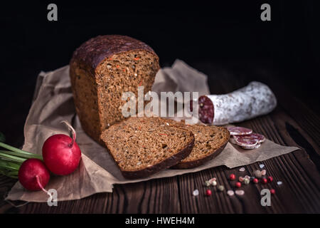 Fette di pane, salumi, ravanello e pepe a secco su carta su sfondo in legno stile rurale vicino fino Foto Stock