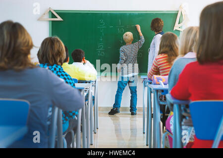 Ragazzo somma di scrittura sulla lavagna nella parte anteriore della classe, vista posteriore Foto Stock