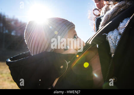 Soleggiato close up baby boy indossando knit hat, eseguito in baby sling da madre Foto Stock