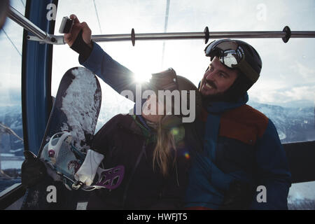 Coppia felice tenendo selfie overhead in funivia contro sky durante il periodo invernale Foto Stock