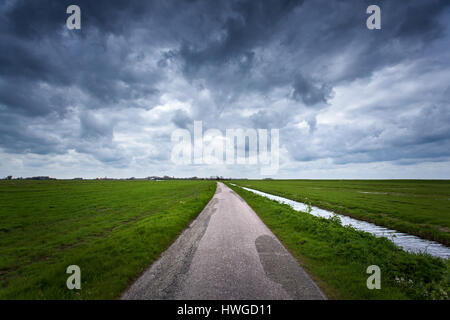 Strada con drammatica azzurro cielo nuvoloso ed erba verde nel freddo giorno di primavera. Fantastico paesaggio nei Paesi Bassi. La natura e la corsa dello sfondo. Strada rurale Foto Stock