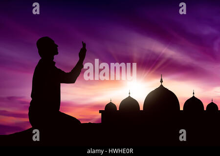 Giovani asiatici uomo musulmano in preghiera a Dio con il tramonto backround e moschea sullo sfondo Foto Stock