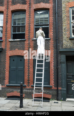 Tradesman commercia l'uomo su una scala che ripara la cornice della finestra di una casa di Huguenot di Londra, Londra orientale, Regno Unito. Lavoratore autonomo Regno Unito. Commerciante Regno Unito. Finestra della scaletta. Foto Stock