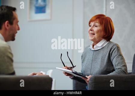 Allegro psicologo maturo con appunti in mani seduto di fronte la sua paziente di sesso maschile e che guardano a lui con sorriso durante la sessione di terapia Foto Stock
