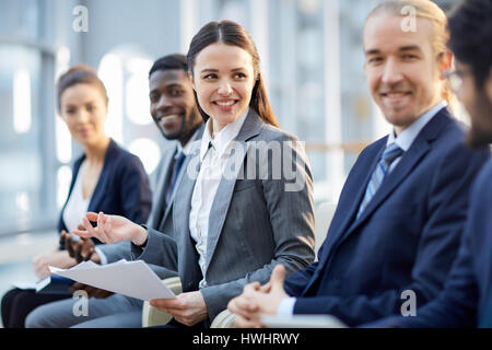 Multi-gruppo etnico di sorridere business persone sedute in fila nella moderna hall di vetro, focus sul giovane bella bruna donna Foto Stock