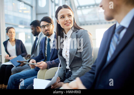 Multi-gruppo etnico di business persone sedute in fila nella moderna hall di vetro, focus sul giovane bella bruna Donna che parla di un irriconoscibile business Foto Stock