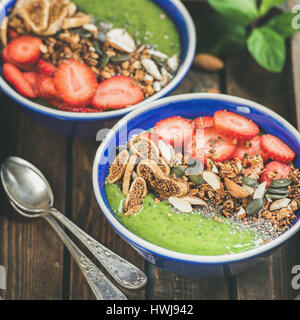 Una sana prima colazione. Frullato verde ciotole con fragole, granola, chia e semi di zucca, la frutta secca e il dado nel vassoio in legno, fuoco selettivo squar Foto Stock