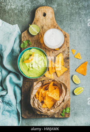 Mais messicano chips, Guacamole fresco salsa in blu ciotola e bicchiere di birra di grano su legno rustico che serve scheda sopra di cemento grigio sfondo tabella, parte superiore Foto Stock