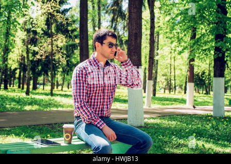 L'uomo in estate nel parco, rilassarsi con una tazza di caffè o di tè, chiamando al telefono con gli occhiali, jeans luminosa giornata di sole. Idea concetto imprenditore prima colazione. Foto Stock