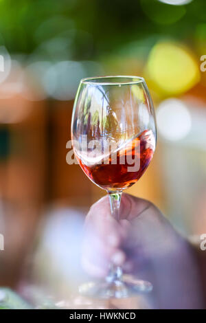 Rosso luce viola il colore rosa vino in un vetro trasparente viene ruotata e ritorto in un bicchiere prima della degustazione. Mano si tiene il vetro di un ristorante esterno Foto Stock