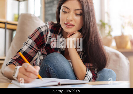 Nuove idee. Felice attraente donna creativa tenendo una matita e notando qualcosa verso il basso mentre si tiene il suo mento Foto Stock