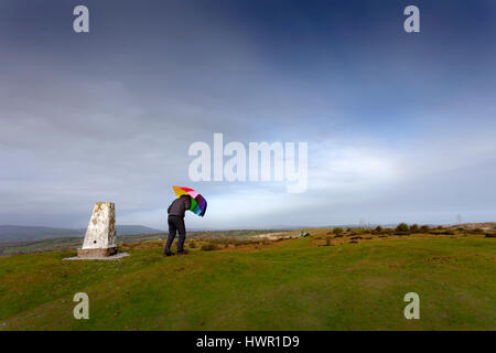 Persona in possesso di ombrello contro il vento accanto ad un pilastro trig sulla Halkyn Montagna in Flintshire, Galles Foto Stock