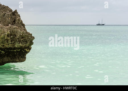 Una linea di riva grande roccia appendere fuori su una luce verde paradiso mare con una grande barca a vela off all'orizzonte. Foto Stock