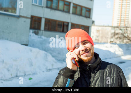 L'uomo emotivamente comunica telefonicamente. guy con la barba, vestito di un autunno sport giacca casual e arancione brillante hat, parla da telefono cellulare in st Foto Stock