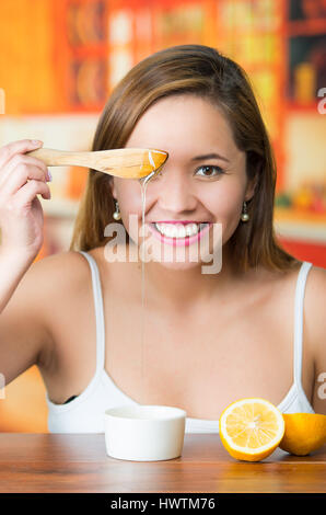 Bruna giovane donna seduta tenendo il coltello in legno con miele dorato il gocciolamento da esso, sorridente felicemente Foto Stock