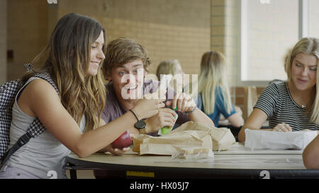 Gli studenti di mangiare il pranzo e utilizzando il telefono cellulare durante la rientranza nella mensa scolastica Foto Stock