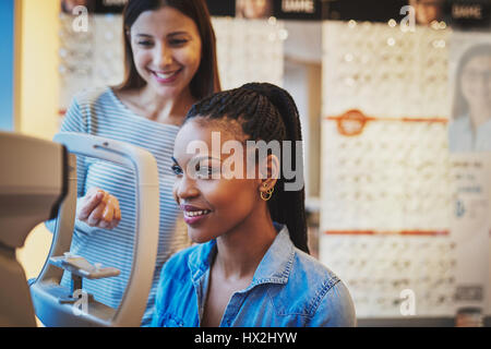 Sorridente giovane nero donna seduta a una macchina in un negozio di ottica è pronto a prendere esame oculistico Foto Stock
