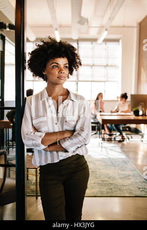 Ritratto di giovane donna africana appoggiata a office porta e guardando lontano. Riflessivo executive femmina in ufficio con le persone che lavorano in background. Foto Stock