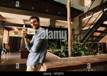 Ritratto di bel giovane uomo in piedi in un ufficio moderno con telefono mobile guardando la fotocamera e sorridente. Business man tenendo pausa dal lavoro. Foto Stock