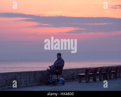 Westbay, Dorset, Regno Unito. Il 24 marzo 2017. Un uomo in un handicap di scooter orologi come il cloud infine si cancella e un vago ma colorato tramonto segue. Con un meteo alto insieme a seguire e temperature più calde previsto per questo fine settimana. © Dan Tucker/Alamy Live News Foto Stock