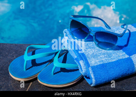 Blue pantofole, occhiali da sole e la salvietta sul bordo di una piscina - vacanza concetto tropicale Foto Stock