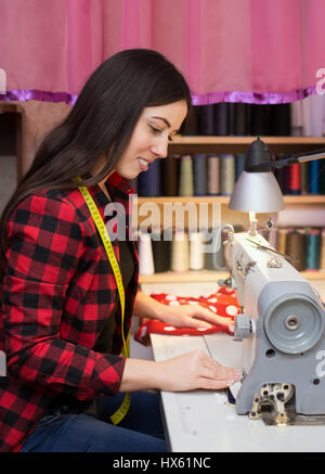 Ritratto di giovane donna seduta sarta e cuce sulla macchina da cucire. Sarta lavorare sulla macchina per cucire. Adattare la realizzazione di un indumento in suo luogo di lavoro Foto Stock