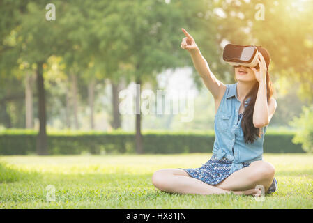 Bella donna asiatica touch screen con la realtà virtuale seduto sull'erba nel parco all'aperto. VR occhiali cuffie dispositivo. natura sfondo all'aperto. peopl Foto Stock