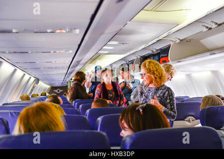 Una vista dal retro di un aereo commerciale della cabina con persone imbarco, seduta & utilizzando contenitori di overhead mentre gli assistenti di volo assistere Foto Stock