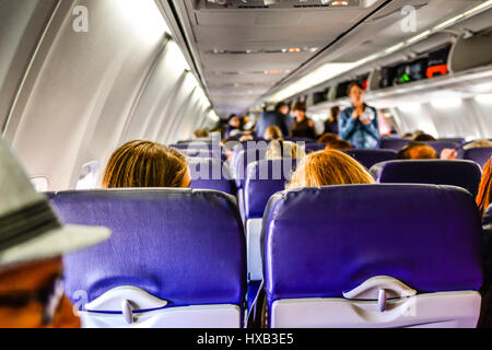 Una vista dal retro di un aereo commerciale della cabina con persone imbarco, seduta & utilizzando contenitori di overhead mentre gli assistenti di volo guarda su Foto Stock