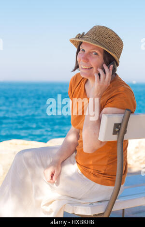 Bella adulto donna caucasica indossando Elegante cappello di paglia è utilizzando il telefono cellulare su Vacanze Estate Mare Vacanze Foto Stock