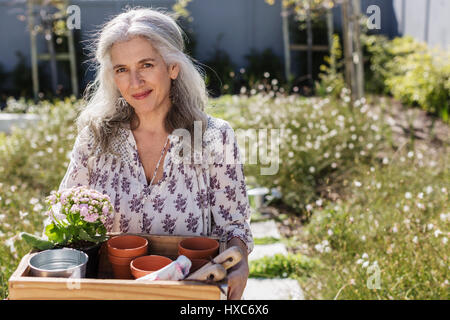 Ritratto di donna matura portante vassoio di giardinaggio nel giardino soleggiato Foto Stock