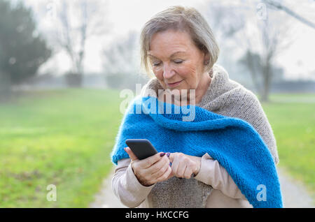 Attraente tech savvy donna anziana utilizzando un telefono cellulare sms di un messaggio a un amico o la navigazione per un numero da chiamare su una corsia rurale in una nebbiosa wi Foto Stock