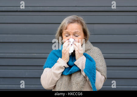 Senior donna in una calda lana lavorata a maglia Sciarpa invernale all'aperto in piedi di fronte a un grigio metallo naso soffiando il suo naso su un tessuto in un concep healthcare Foto Stock