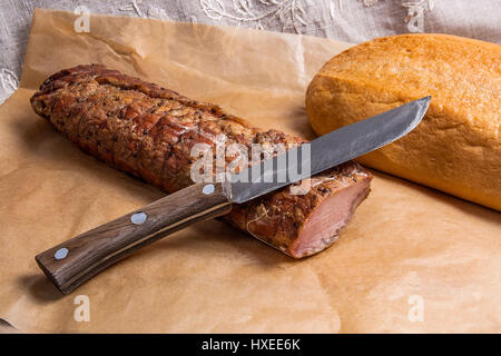 Affumicato con spezie e erbe aromatiche a base di carne o prosciutto, vecchio coltello e bianco pane di frumento su marrone carta da imballo. Panno marrone sul retro dello sfondo. La composizione della luce Foto Stock