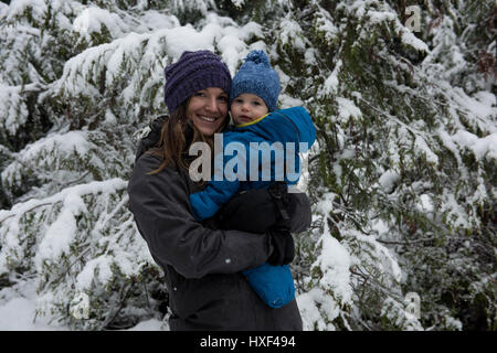 Ritratto di Madre sorridente che trasportano figlio contro la coperta di neve alberi Foto Stock