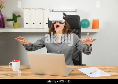 Business donna, con camicia grigia seduta, guardando con la realtà virtuale la cuffia o 360 bicchieri, interagendo con amazed felice espressione, e portatile Foto Stock