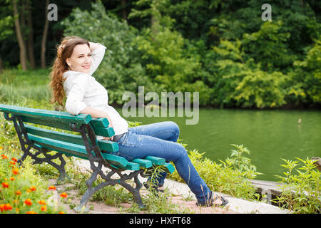 Giovane donna in maglia bianca seduta sul banco vicino al laghetto nel parco Foto Stock