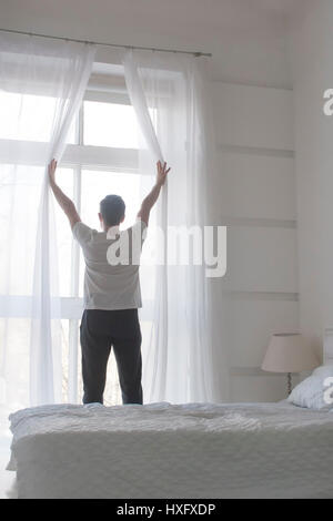 Giovane uomo di aprire le tende per welcomw mattina e luce, vista posteriore, orientamento verticale Foto Stock
