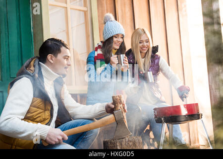 Giovane uomo trinciatura di legna da ardere con ax mentre felici donne bere il tè vicino grill Foto Stock