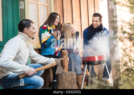 Uomo sorridente trinciatura di legna da ardere con ax mentre happy amici bere il tè vicino grill Foto Stock