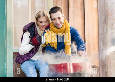 Felice coppia giovane in vestiti caldi seduti insieme vicino grill sul portico Foto Stock