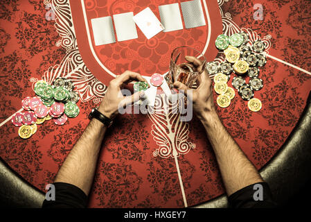 Vista parziale di uomo seduto al tavolo del poker con bicchiere di drink alcolici e poker chip in mani Foto Stock