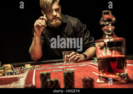 Bel uomo barbuto di fumare un buon sigaro e bere whisky giocando a poker Foto Stock