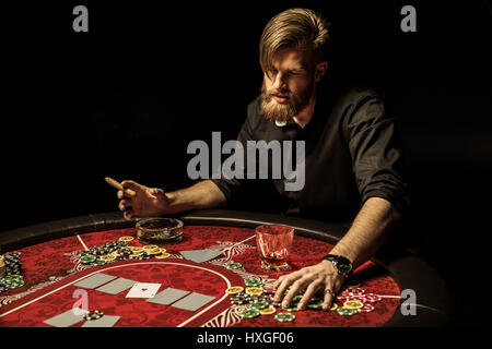 Bel uomo barbuto di fumare il sigaro giocando a poker a tavola Foto Stock