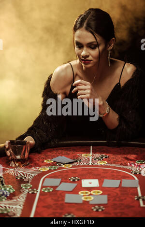 Stupendo giovane donna in abito da sera seduti al tavolo del poker con un bicchiere di whisky Foto Stock