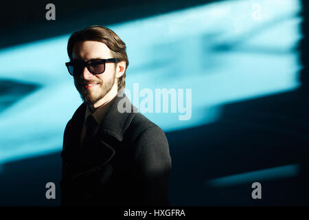 Ritratto di uomo bello indossare occhiali da sole e mantello nero in condizioni di luce solare intensa contro sfondo blu e sorridente splendidamente guardando la fotocamera Foto Stock