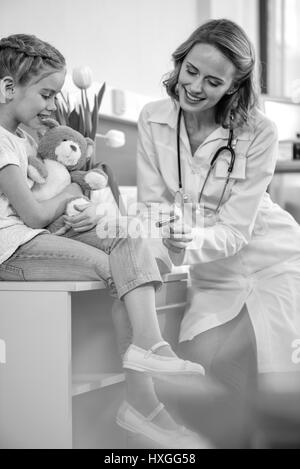 Vista laterale del medico sorridente esaminando ragazza con martello di riflesso, foto in bianco e nero Foto Stock