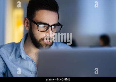 Ritratto di bello uomo barbuto con gli occhiali a lavorare con il computer portatile in ufficio scuro a tarda notte, il suo viso illuminato dallo schermo Foto Stock